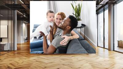 Loving family of three at home sits on the comfortable sofa. Multiracial parents hold biracial newborn baby and look at her with love and care Wall mural