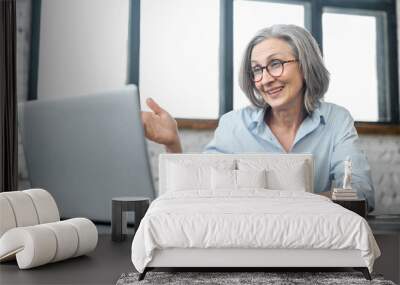 Mature aged business woman using a laptop for online video meeting with a colleagues. Senior teacher conducts classes, webinars. An elderly lady sits at the desk and talking at the online conference  Wall mural