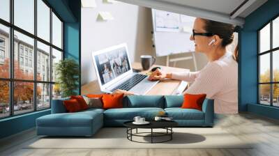 Morning meeting online. A young woman is using app on laptop for connection with colleagues, employees. Video call with many people together. Back view Wall mural