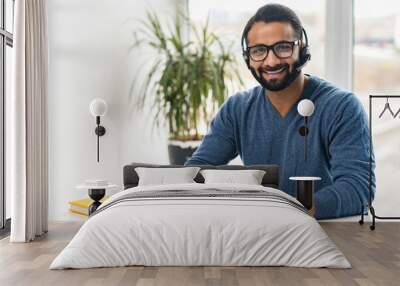 Portrait of cheerful Indian guy in wireless headset using laptop computer sitting in contemporary loft style office space, Hispanic man is making video call, talking online on distance Wall mural