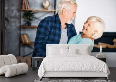 Portrait of modern romantic elderly couple is hugging, embracing gently, dancing at home. Senior spouses stand holding hands, looks to each other and laugh happily Wall mural