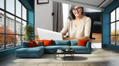 Senior business woman using a laptop for remote work from home, confident mature lady checking emails, conducts business correspondence sitting at the table, looks at the camera and smiles Wall mural