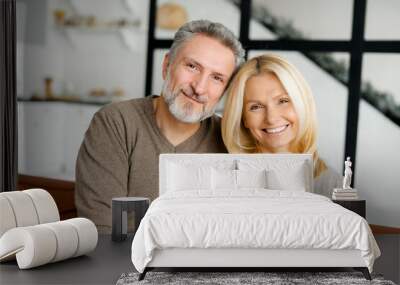 Serene and carefree middle-aged couple in love looks at the camera and smiles. Good-looking grey-haired handsome man and charming blonde woman sit in embrace on the couch. Love and bonding concept Wall mural