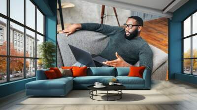 Serious African American guy with glasses lies on the couch and using laptop for online talking via video call. A black guy in glasses looks at laptop screen and talks emotionally Wall mural