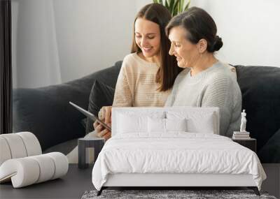 Side view an adult daughter and a senior mother spend time with a tablet indoor, a young woman showing to mature mom smth in a network, helps to figure out with a device Wall mural