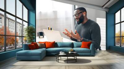 Side view of African-American office employee talking via video connection while sitting at the desk. A guy using laptop for video call in contemporary office space Wall mural