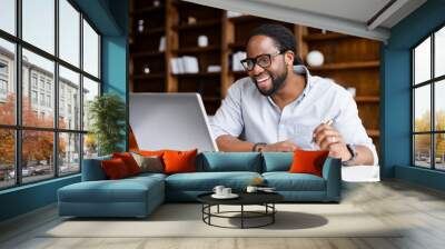 Smiling African-American guy takes notes watching webinars on the laptop,researching business tasks, a male student is studying online, listening video lectures, writes down, e-learning concept Wall mural