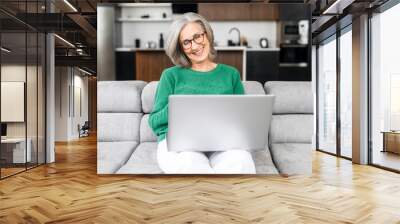 Smiling charming elderly woman looks at the screen of the laptop and laughs sitting on a couch at home, an elderly lady wearing eyeglasses spends leisure in networks, watching movies, has a video call Wall mural