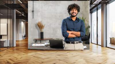 Smiling curly middle eastern businessman standing near desk and looking at the camera. Young positive male student with modern flat on background. Proud and successful mixed-race small business owner Wall mural