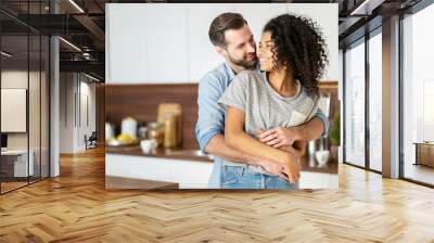 Smiling man hugging from behind charming African American woman, two people standing and joyfully looking at each other. Young international couple happily spending time in cozy modern kitchen at home Wall mural