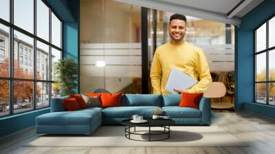 Smiling young indian businessman, arab male student, eastern employee, manager, coworker wearing casual yellow pullover standing carrying laptop, looking at the camera standing in office Wall mural