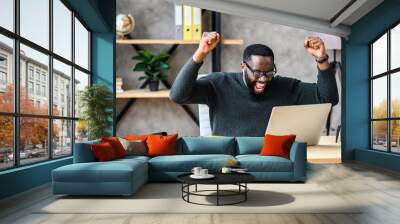 Success at work, good deal. African-American guy looks at the laptop screen, screams excitedly and raises his fists in a victory gesture Wall mural