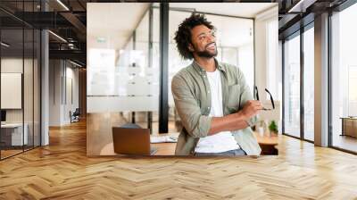 Successful cheerful african-american male entrepreneur, small business owner, male office employee, black businessman wearing green casual shirt stands in confident pose with arms crossed and laughing Wall mural