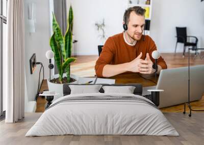 The guy uses hands-free headsets to work from home. He sits at a table with a laptop and speaks online. Remote work concept Wall mural