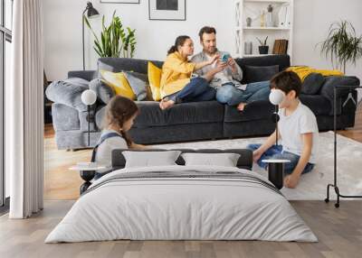 Two parents using smartphones sitting on the sofa in cozy living room, scrolling news feed while two kids playing with constructor on the floor, sibling enjoying game together. Family of four at home Wall mural