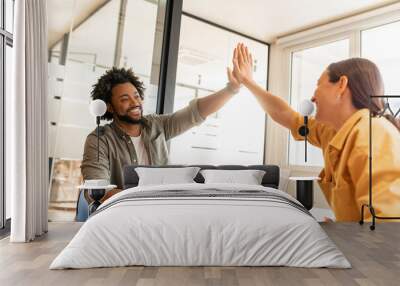 Two young happy diverse colleague sitting at desk and taking high five while working together in coworking space, man and woman celebrating successful project completion or good deal Wall mural