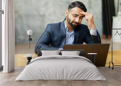 Upset Indian businessman in formal suit has strong headache, sitting in front of laptop and holding forehead, feels tired and exhausted, sad eastern man worrying, feels anxiety, suffers from migraine Wall mural