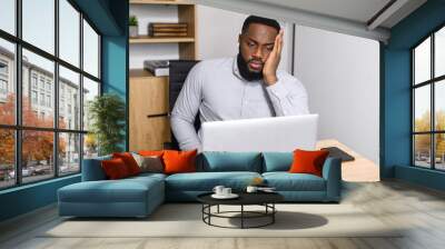 Young African-American male worker feeling stressed, tired, and overworked, holding head in hands, sitting at the desk in front of a laptop, applying problem-solving skills. Deadline struggle concept Wall mural