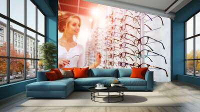 Row of glasses at an opticians. Eyeglasses shop. Stand with glasses in the store of optics. Woman chooses spectacles. Eyesight correction. Wall mural