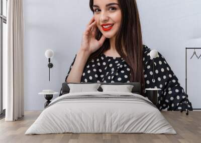 Young woman with long hair smiling in polka dot dress. A young woman with long brown hair smiles confidently, wearing a stylish black and white polka dot dress against a light background. Wall mural