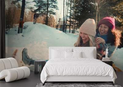 Two attractive multiethnic happy women smile sledging on snow slope towards camera, winter weekend fun slow motion. Wall mural