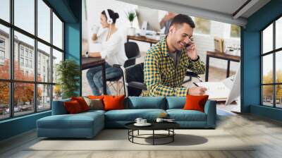 A man in a wheelchair writes with a pen in a notebook and talks on the phone. He is working in a bright office. Wall mural