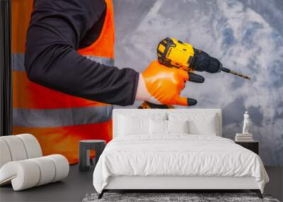 Male worker holds a close-up electric cordless screwdriver in his hands against the background of a construction tool and a concrete wall. Wall mural