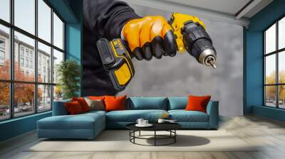 Male worker holds a close-up electric cordless screwdriver in his hands against the background of a construction tool and a concrete wall. Wall mural