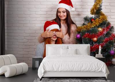 Young beautiful girl closing her friend's eyes for a surprise. Two women celebrating Christmas in Santa caps sharing gifts near Christmas tree. New Year, Christmas time, surprise, celebration concept Wall mural