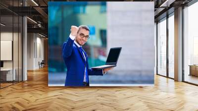 Young happy businessman standing on office building background with laptop in hand and expressing positivity, copy space Wall mural