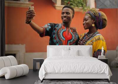 African young stylish and attractive couple in traditional outfits standing together outside and posing to the smartphone camera while taking selfie photo. Wall mural