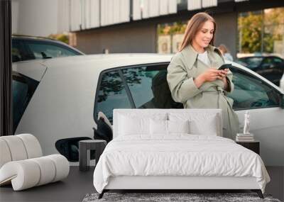 Beautiful young woman smiling standing near white electric car while charging her auto outdoor and texting on mobile phone. Caucasian casual female browsing on smartphone in city at EV charger station Wall mural