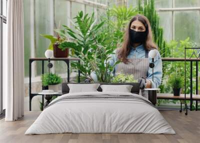 Caucasian beautiful happy woman entrepreneur owner of floral shop in black mask holding floral box in hands indoors. Pretty young female employee in garden center with flowers and plants. Job concept Wall mural