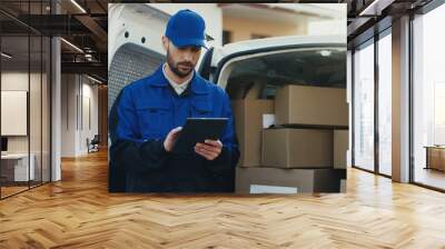 Caucasian good looking mailman in blue costume and cap checking the mail boxes with tablet device in hands. Outdoor. Camera zooming in. Wall mural