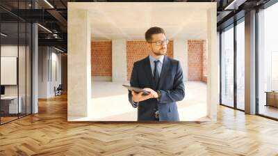 Caucasian male real-estate agent in glasses, suit and tie walking in building on construction phase, using tablet device and looking around. Businessman choosing mortgage. Wall mural