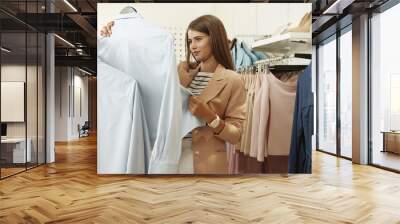 Cheerful pretty young Caucasian female buyer in fancy clothing shop looking at clothes and taking from hanger. Charming girl choosing outfits in store. Woman shopper buying wear. Wall mural