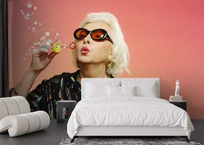 Close up of old fancy and beautiful Caucasian woman with gray hair and in sunglasses blowing soap bubbles on pink wall background. Pretty grandmother in stylish outfit playing. Wall mural