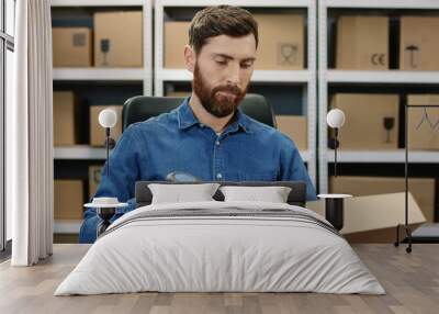 Handsome Caucasian postman sitting at desk and working at computer in postal delivery store. Man mail worker scanning parcel and registering it in system in post office. Wall mural