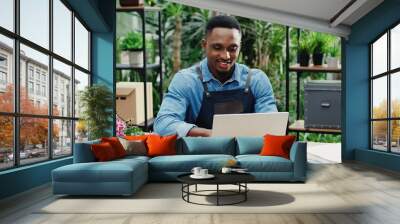 Joyful young man in flower shop sitting at workplace and texting on computer. Cheerful African American male florist worker in floral house tapping and typing on laptop. Floristry concept Wall mural