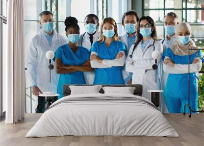 Mixed-races busy team of male and female doctors posing to camera and crossing hands in hospital. International group of medics in medical masks. Protected multi ethnic physicians and nurses in clinic Wall mural