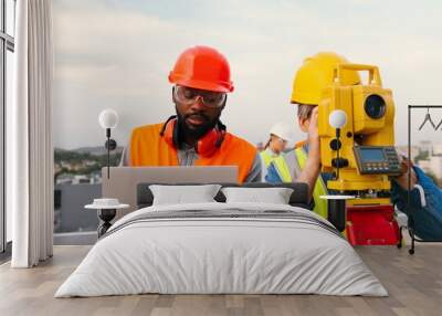 Mixed-races men constructors at building site working and measuring angle with total station on roof. Multi-ethnic males topographers. Caucasian and African American builders with laptop computer. Wall mural