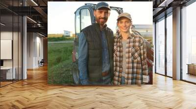 Portrait Caucasian happy couple of married farmers standing in hugs at tractor, looking at each other and smiling to camera. Outdoor. Man hugging woman in field. Agriculture concept. Wife and husband. Wall mural