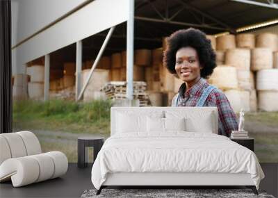 Portrait of beautiful African American young woman farmer standing in field at shed with hay stocks and smiling to camera. Pretty female with smile at farm. Countryside concept. Wall mural