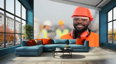 Portrait of handsome African American young man in casque, goggles and headphones standing at construction site and smiling to camera. Happy cheerful male builder at building outdoors. Wall mural