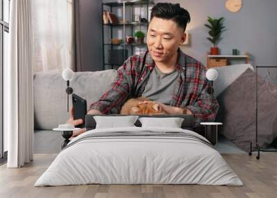 Portrait of handsome caring Asian man sitting on sofa in cozy living room and stroking lovely fluffy cat with hand while reading book. Relaxed cute animal pet in owner's arms, pet lover, leisure hobby Wall mural