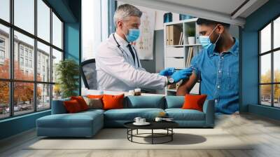 Portrait of handsome middle-aged male professional physician make covid-19 vaccine injection to young Hindu man patient in medical mask sitting in hospital room, coronavirus vaccination in quarantine Wall mural