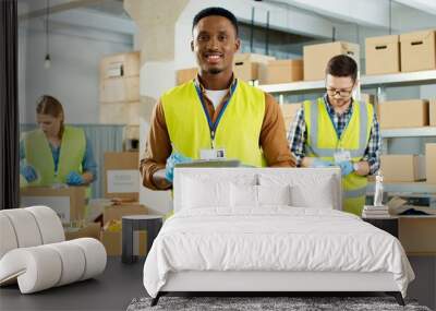 Portrait of happy young African American male social worker standing in storage house checking donation list and smiling to camera. Caucasian volunteers packing food and stuff in boxes charity concept Wall mural