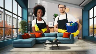 Portrait of joyful young multi-ethnic male and female workers standing in room in apartment in aprons looking at camera and smiling holding cleaning tools, cleaning service concept, small business Wall mural