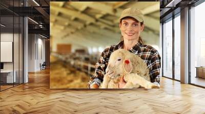 Portrait of young beautiful Caucasian woman farmer in cap smiling to camera and holding lamb in hands in barn with livestock. Happy female shepherd caressing cute animal in stable. Petting sheep. Wall mural