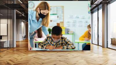 Portrait of young beautiful Caucasian woman teacher helping little African American junior student boy to write test in copybook at class. Female teaching bright schoolboy explaining lesson Post-covid Wall mural